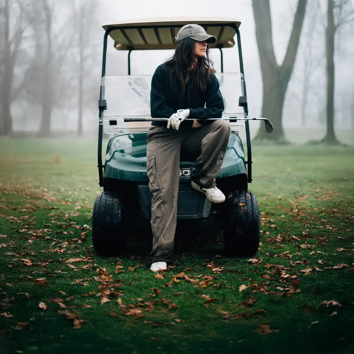 Woman wearing a cap and golf gloves sitting on a golf cart holding a golf iron, in a foggy golf course with autumn leaves.